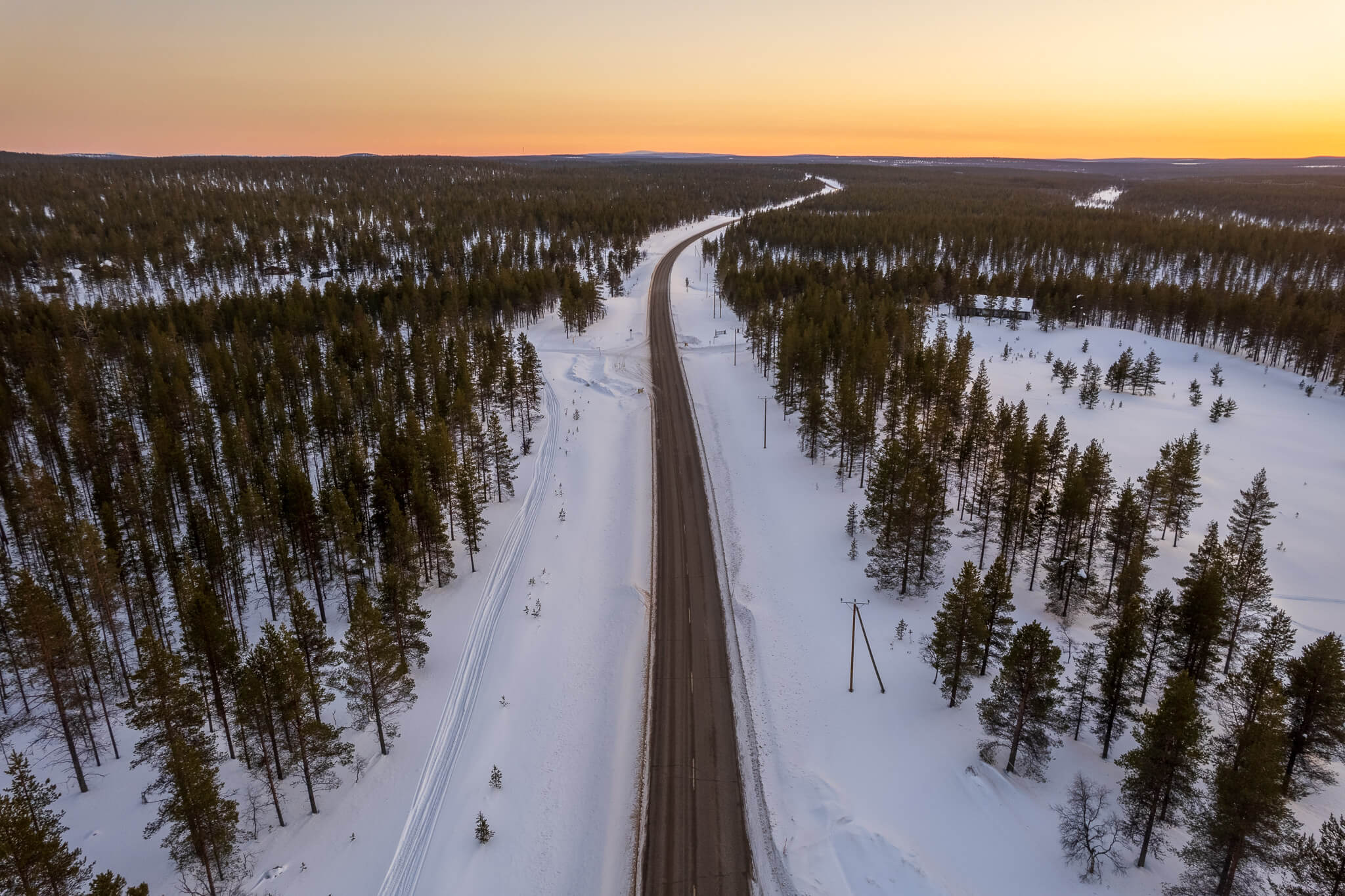 Aerial view of straight road cutting through snow forest at pink sunset, Lapland