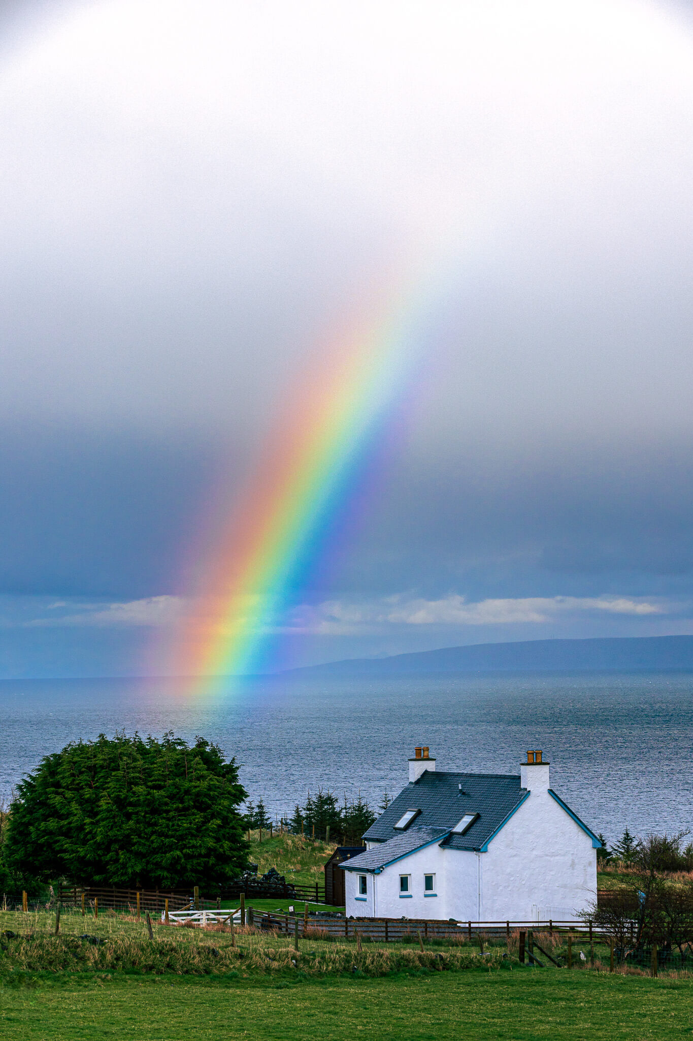 Vivid rainbow arcing over white Scottish cottage beside the sea, Isle of Skye