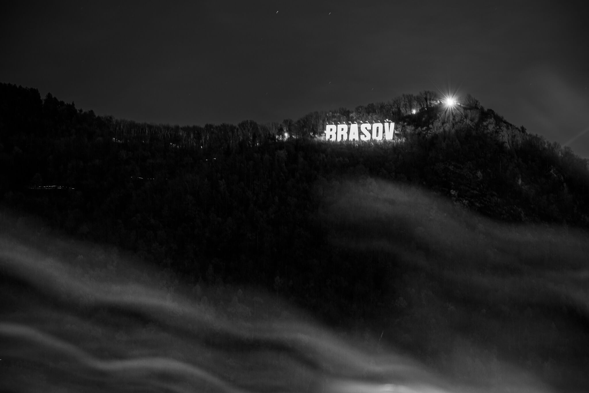 Black and white long exposure of illuminated Brasov sign on hillside above fog, Romania
